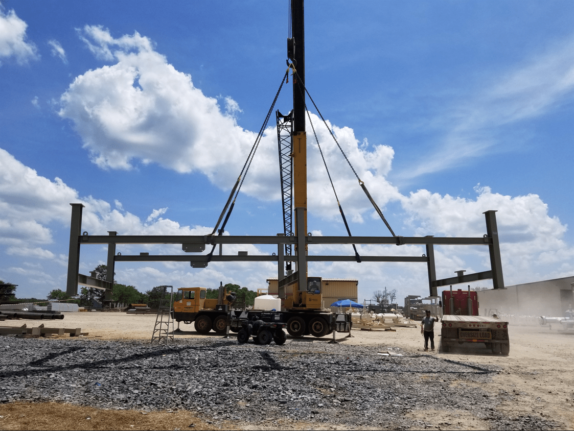 Crane lifting a large industrial frame on a construction site under a blue sky with clouds