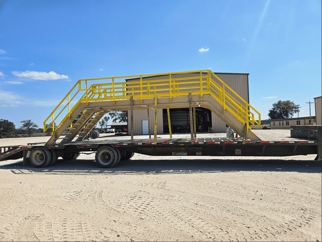 Large industrial access platform with yellow safety rails loaded on a trailer, in a yard on a sunny day