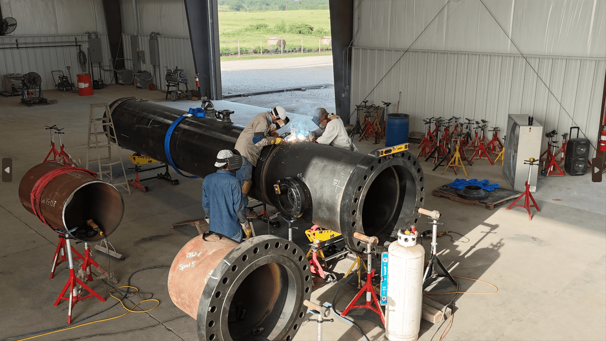 Welders assembling a large industrial pipe section in a workshop with various equipment and supports