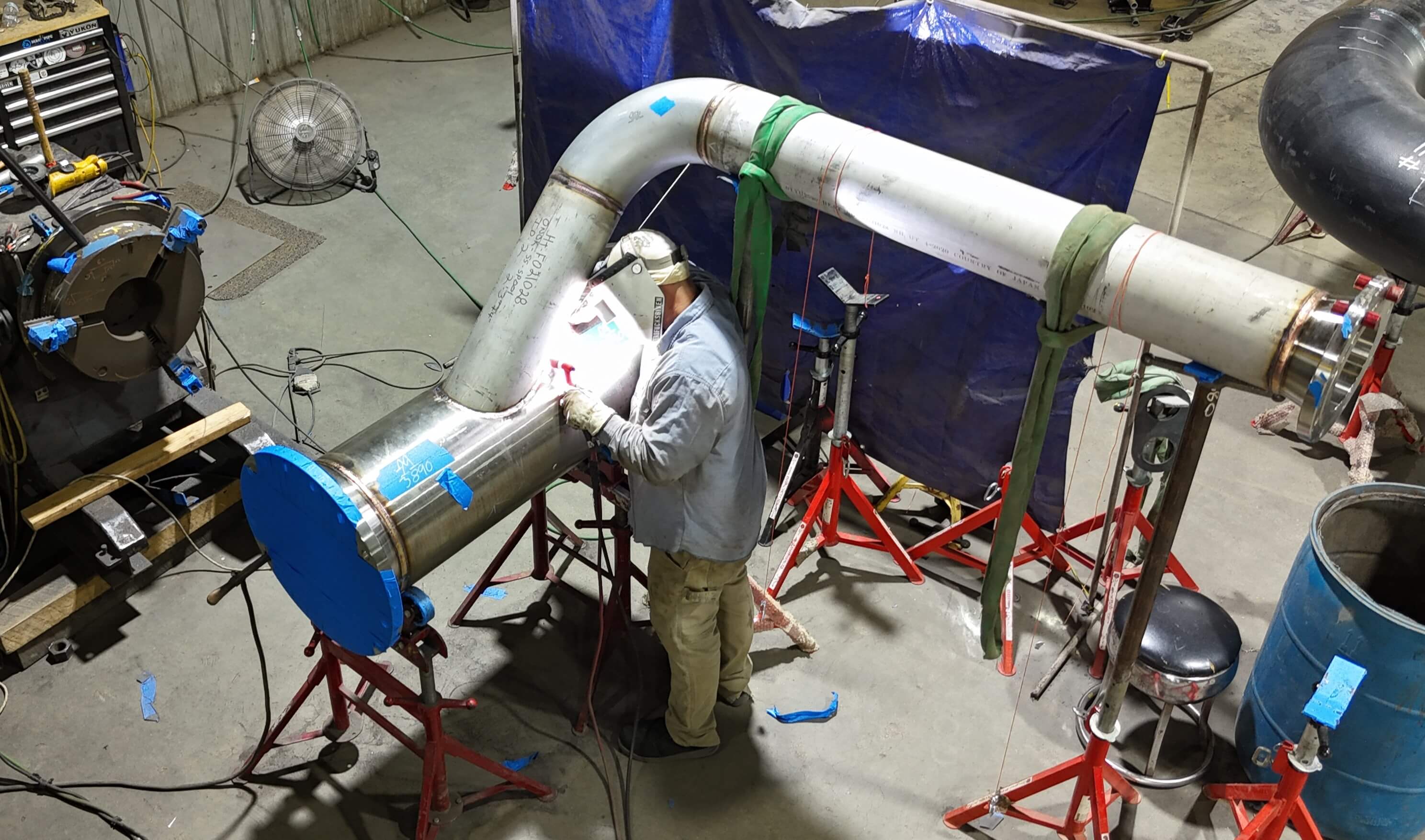 Welder working on a large industrial pipe assembly in a workshop, surrounded by equipment and supports