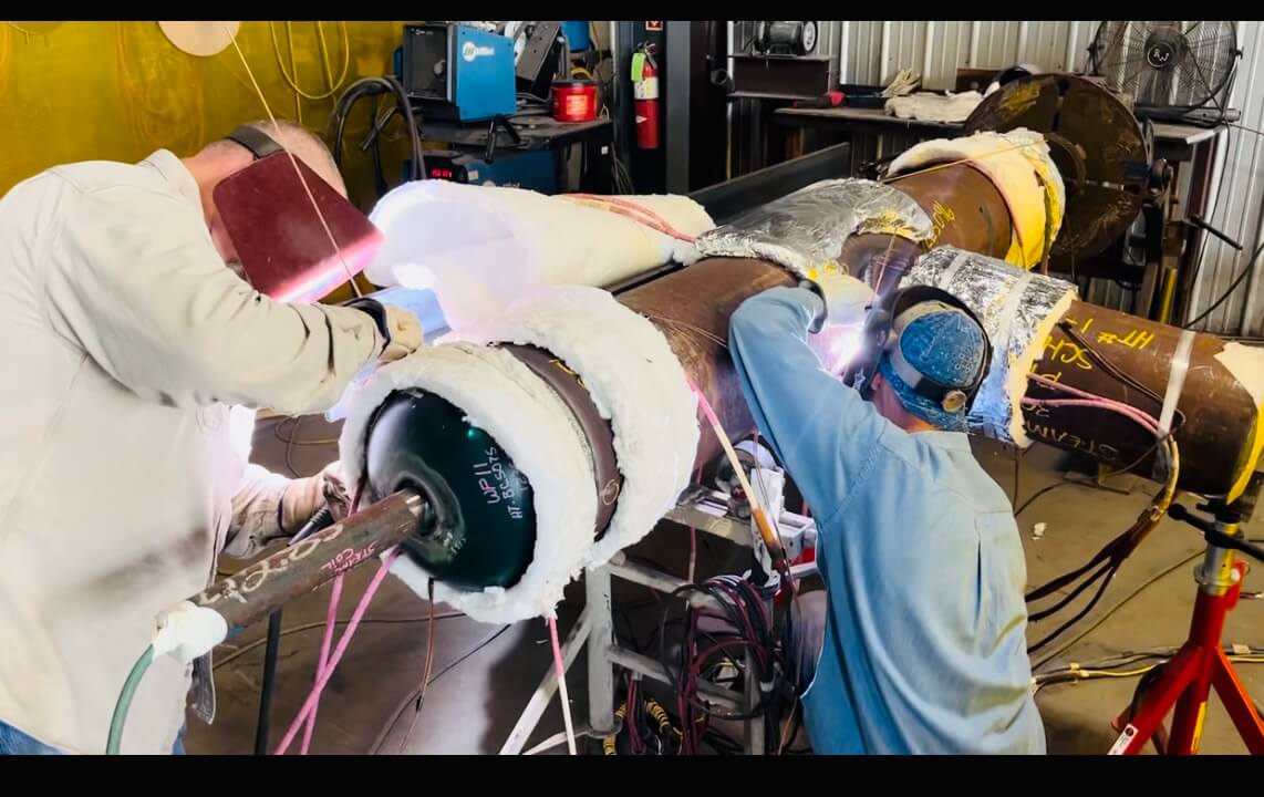 Two welders working on an insulated industrial pipe, surrounded by protective coverings and workshop equipment