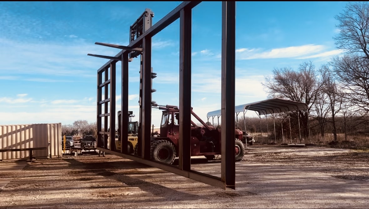 Forklift moving a large steel frame at a construction site, with open sky and trees in the background