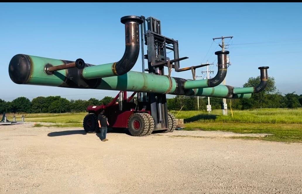 Forklift transporting a large industrial pipe with multiple flanges and fittings in an open yard