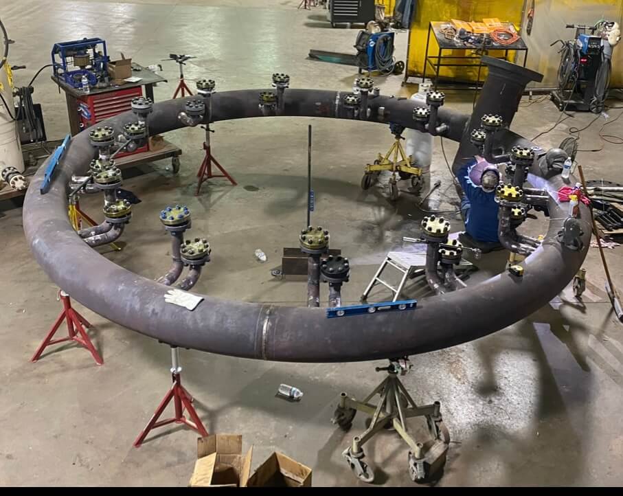 Circular industrial pipe assembly with multiple flanges in a workshop, supported by stands and surrounded by equipment