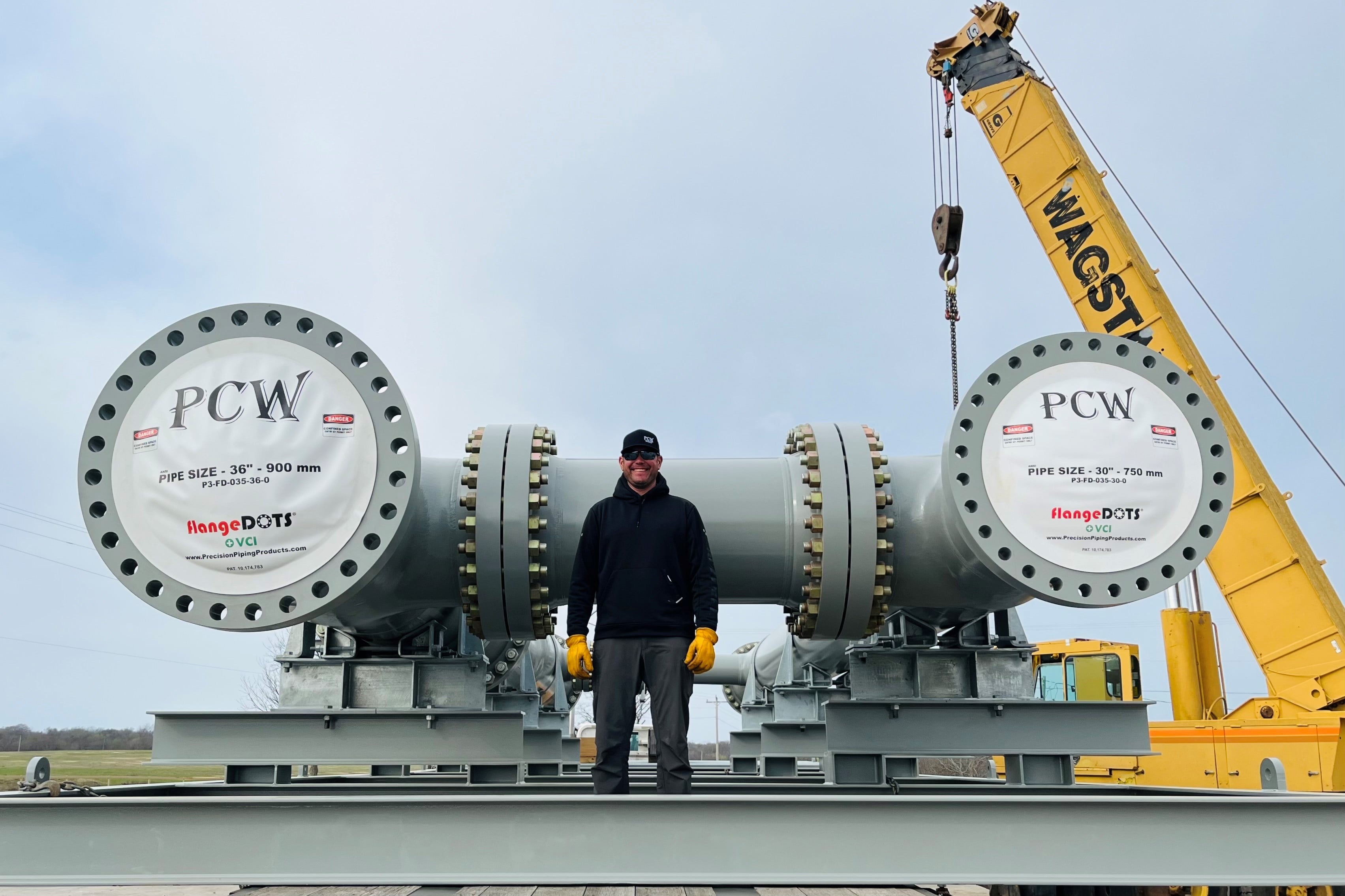 A man standing between large industrial pipes labeled 'PCW', with a crane in the background